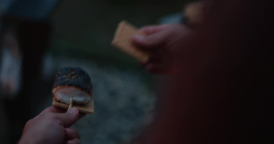 A man is holding a hot roasted marshmallow on a stick as he smashes it between two graham crackers