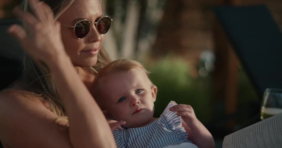 Blonde mother with sunglasses holds baby while leafing through a book