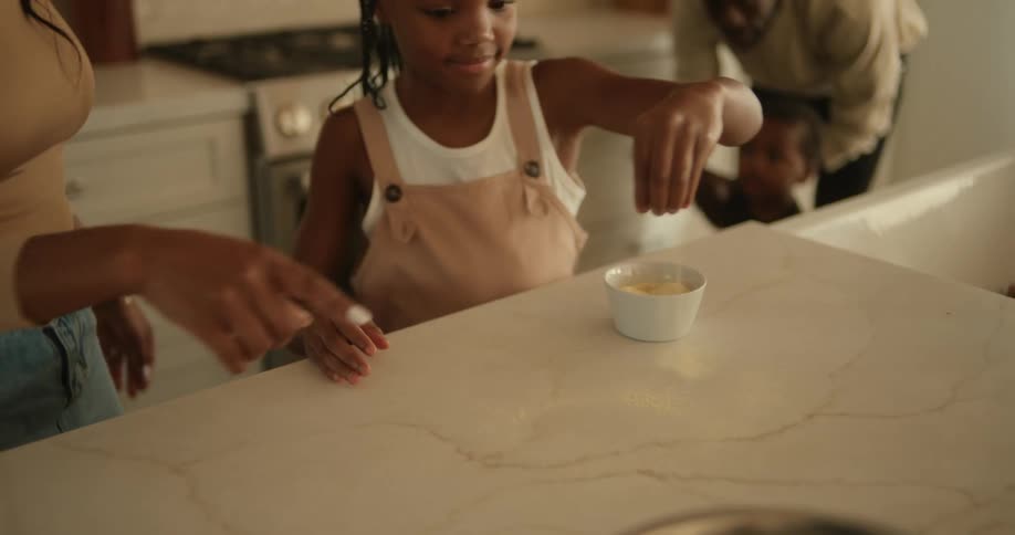A family are in the kitchen as a little girl spreads cornmeal from a cup across the counter as her mother stands next to her