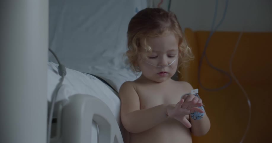 A little girl is standing in a hospital room with an oxygen tube across her face and a nasal cannula in her nose as she plays with her hand