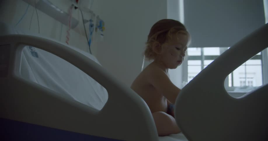 A little girl is sitting on a hospital bed playing as she stops and looks up to her side