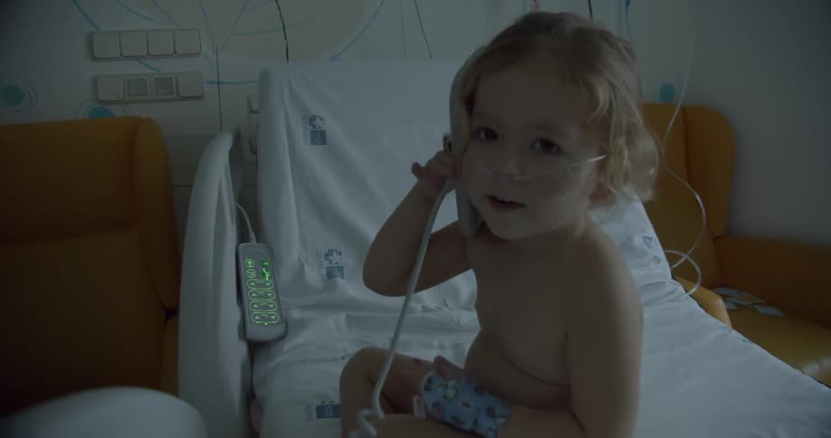 A young girl in a hospital bed with a breathing tube in her nose holding a device to her ear