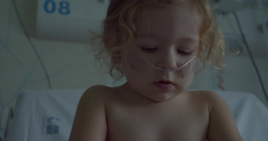 Female toddler indoors sitting down on a hospital bed with a nasal cannula next to a wall with hospital machines and devices during day time