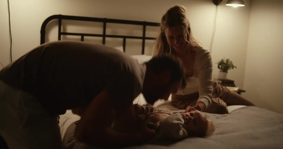 The dad dresses the baby who is lying on the bed while the mom sits beside them