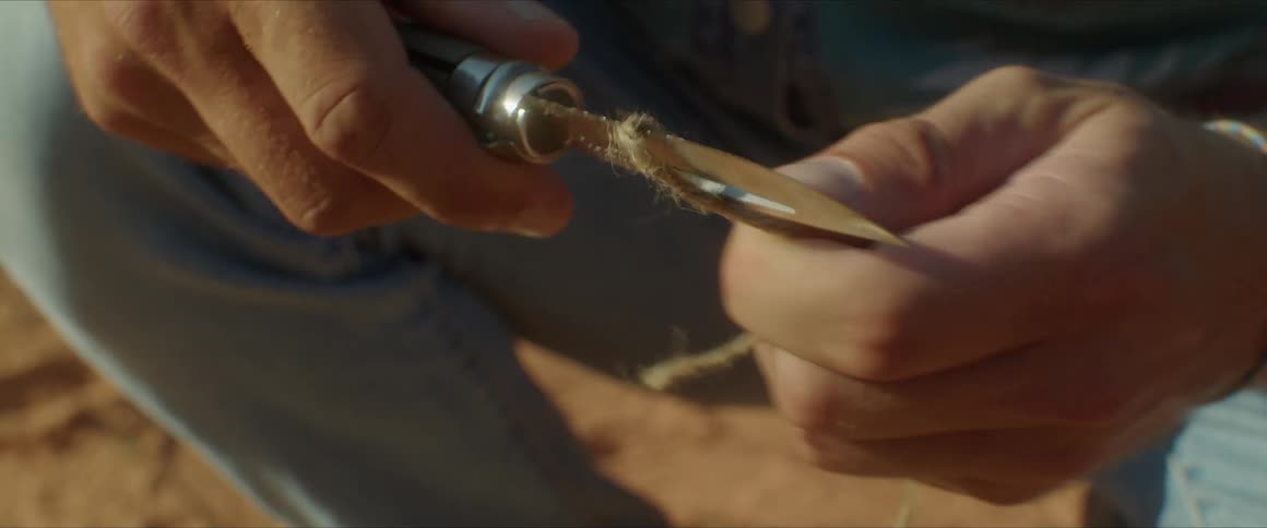 A man wearing jean squat on a dry sand as he cut through a thin rope with a small knife