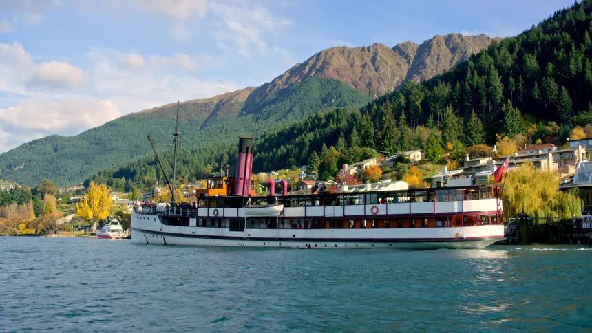 A steamship filled with people cruises across a lake near forested mountains and trees in fall colors near houses in New Zealand