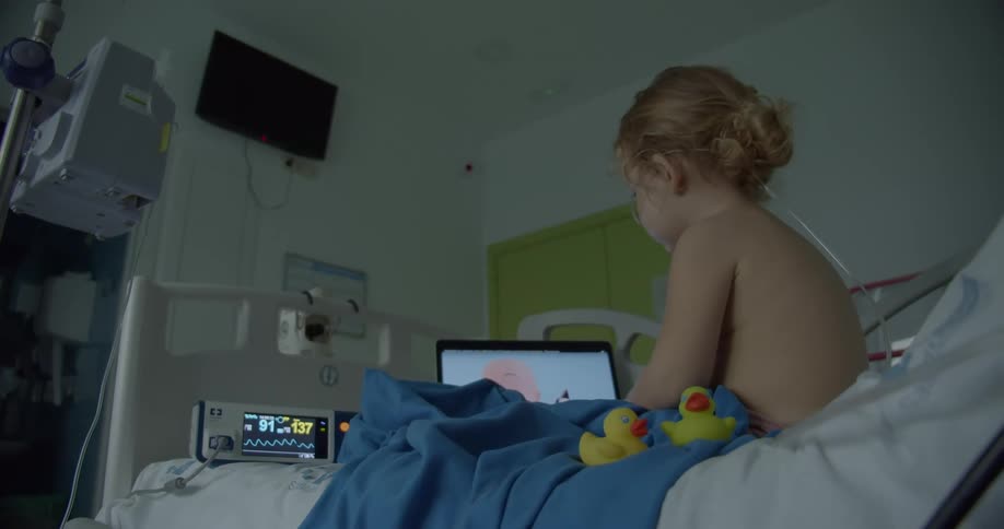 A small blonde child is sitting in a hospital bed with various equipment while watching something on a screen
