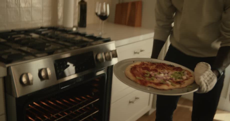 A man taking a pizza out of an oven and placing it on the table while a baby on someones lap watches him