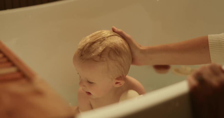 A woman bathting her son on the bath tub while smiling as she pours water with a small cup over the head of the child as he plays around
