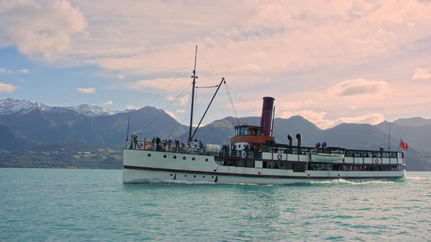 A steamboat with crew members navigating the lake with a mountain range in the background