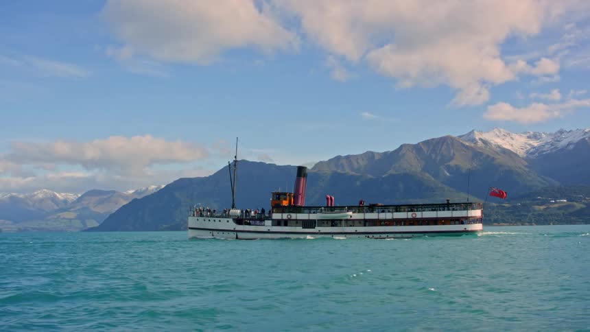 A steamship filled with people cruises across a lake with mountains along the coast under a cloudy sky