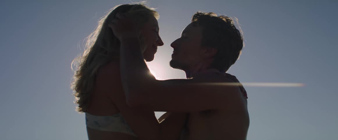 A shirtless man and woman kissing under the sunset