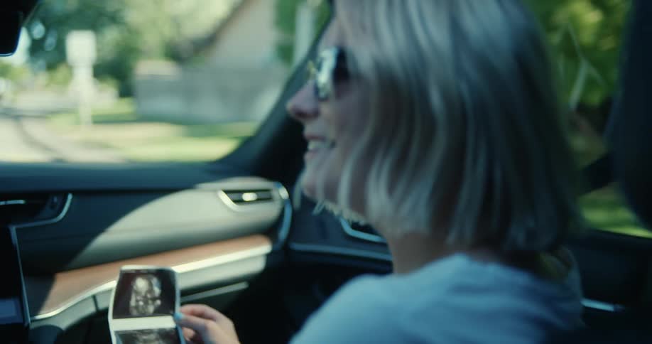 A blonde woman with a photo in her hand inside a car and smiling