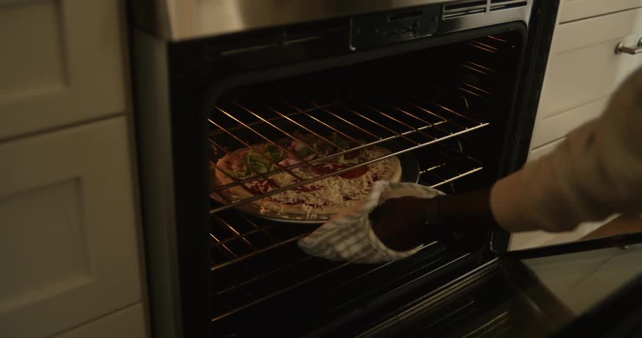 The father slides a pizza into the oven with his daughter nearby as the mother watches from the behind