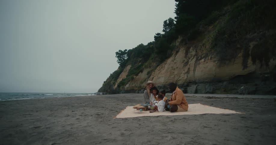 A man and his wife with his two kids sitting on a mat in the beach having a picnic