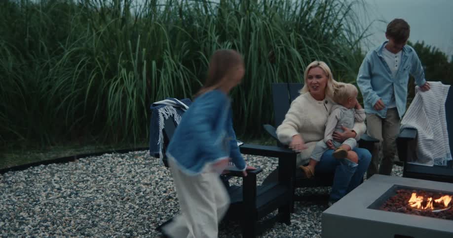 Parents sitting around a campfire roasting marshmallows as their children arrive and take seats in chairs around the fire