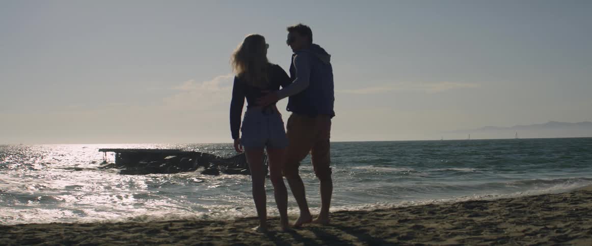 Man and woman cuddling on the beach near the ocean are silhouetted against the sun
