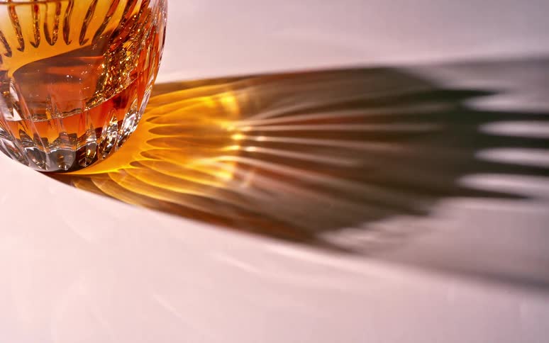 A golden liquid in a glass making a shadow on a white surface that moves as the light passes