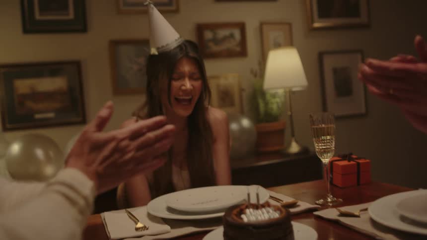 A woman blowing out the candles on her birthday cake with two people sitting and clapping