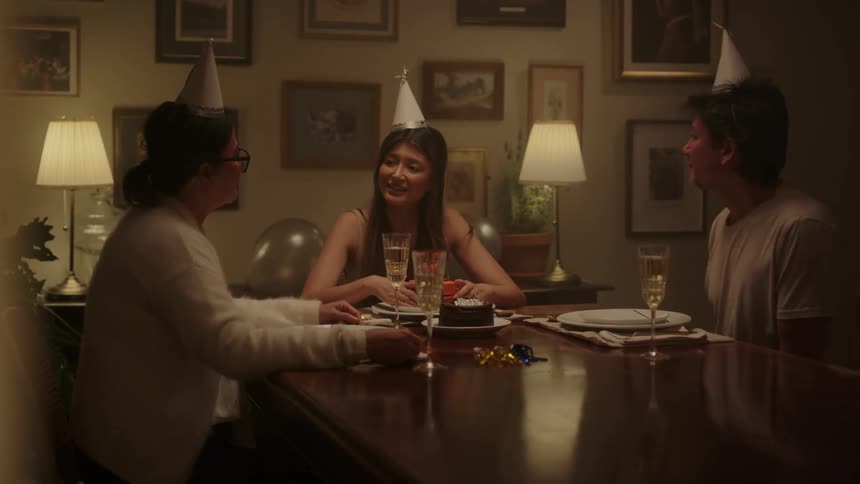 A woman and two men sitting in the dining at home discussing and smiling at each other while wearing a birthday hat