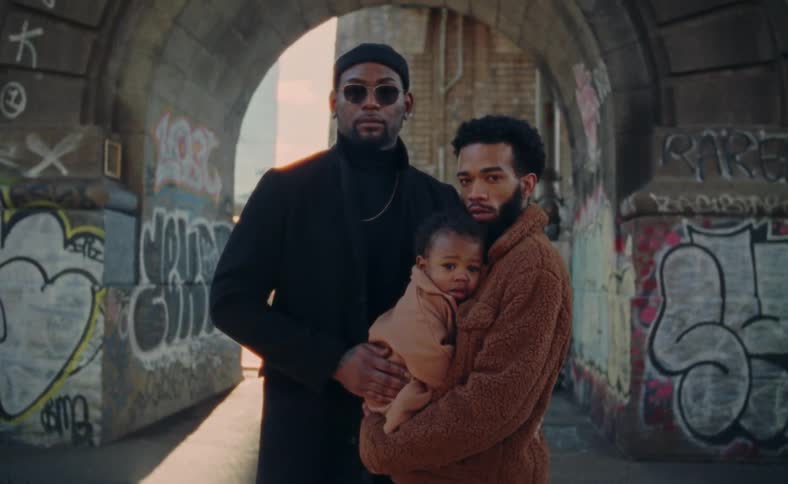 Two black men stand together holding their baby by graffiti outdoors