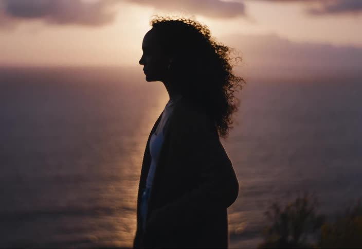 Woman stands silhouetted against the setting sun perched atop a cliff as she gazes out to the ocean