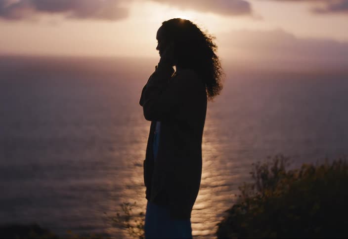 A silhouette of a woman standing sideways to an ocean being illuminated by sunlight