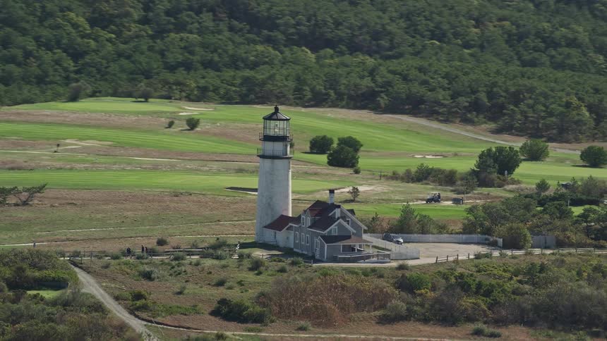A house is next to a lighthouse as people walk on the road in a rural neighborhood surrounded by forests