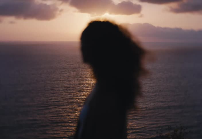 Woman standing on shore on windy day is silhouetted by sun as paraglider flies through the sky in the background