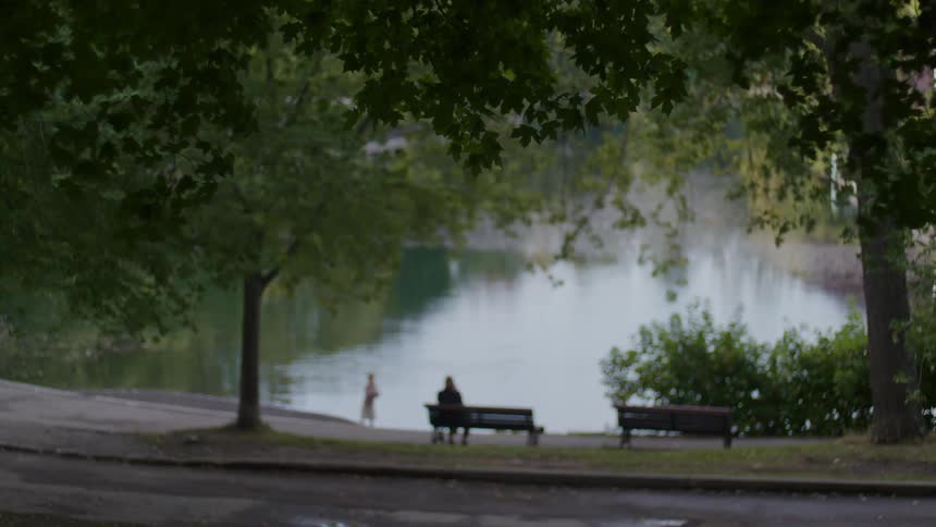 An adult sitting on a bench close to a river as a child plays by the river bank
