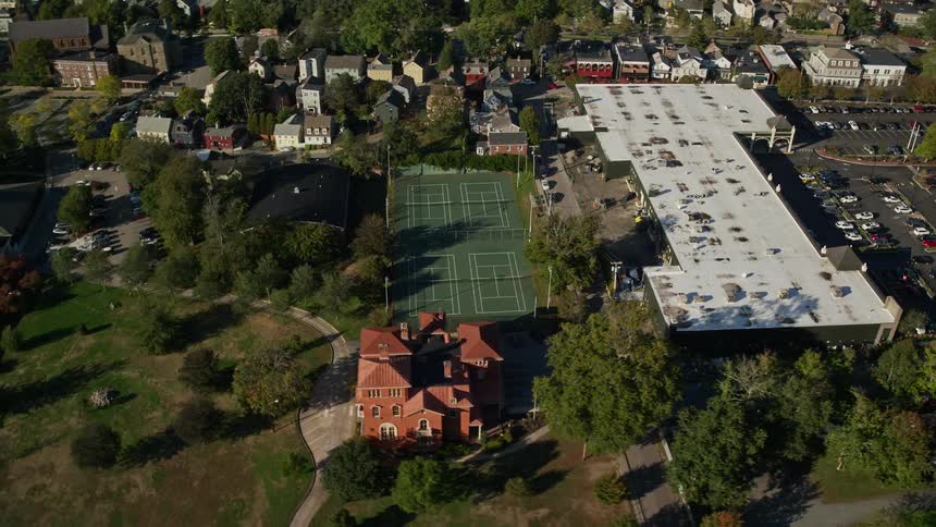 A tennis court is close to some buildings and several trees