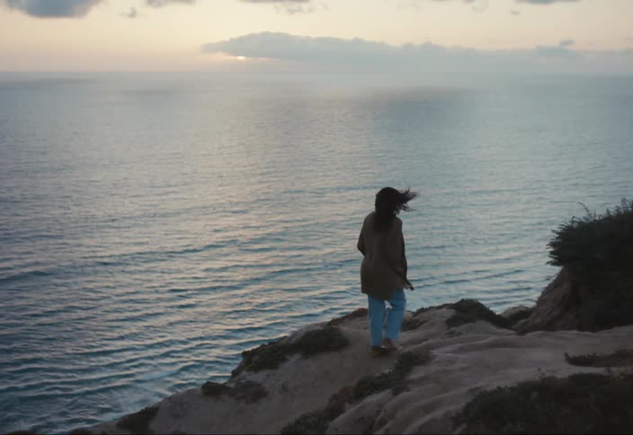 A woman walks along the coast as waves roll in under a hazy cloudy sky