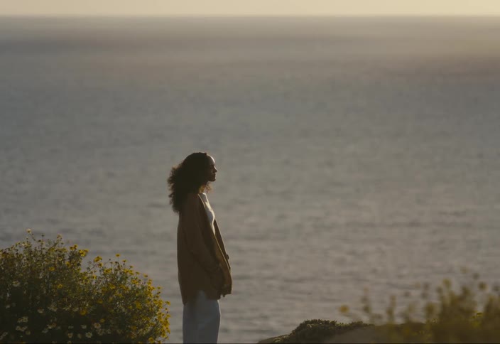 Woman stands on coast among flowers on a windy day and looks at the view