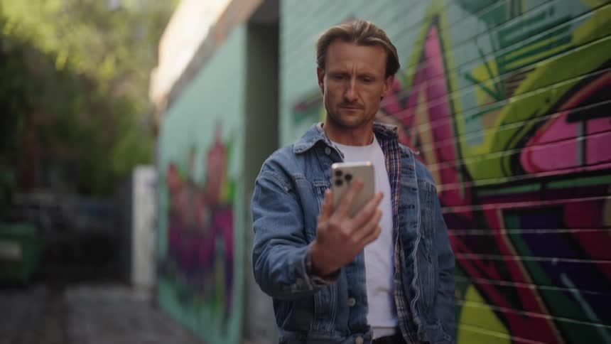 A man is looking at his phone as he walks next to a building with a colorful mural