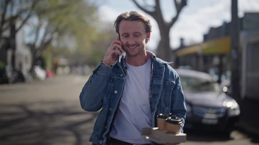 A man is walking down a neighborhood street carrying a tray with two cups of coffee and answers his phone and talks as he walks