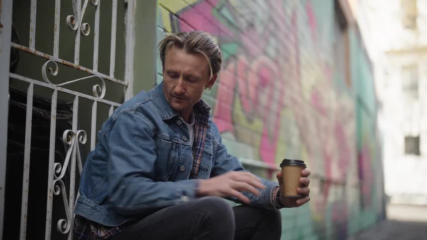 A man is sitting near a building as he drinks a cup of coffee and answers his phone