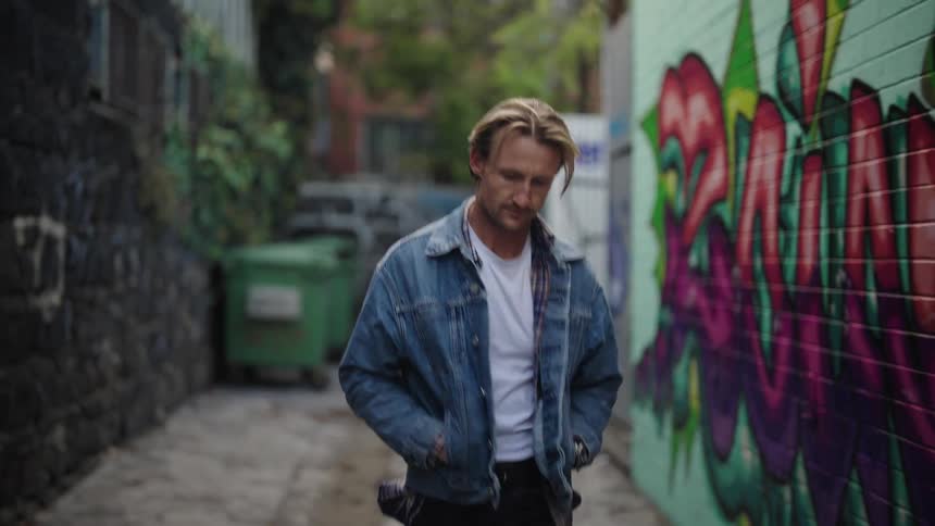A man wearing a denim jacket walks between buildings with his hands in his pockets