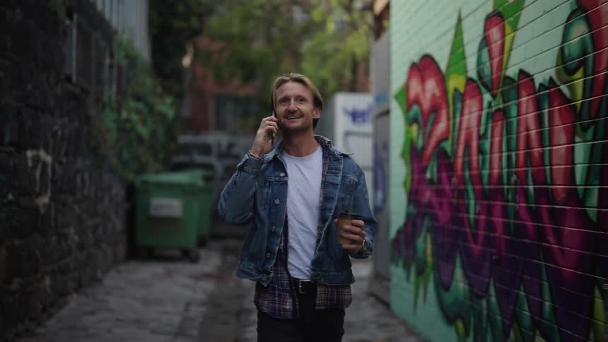 A blond man walking and drinking from a glass while talking to someone on a cell phone