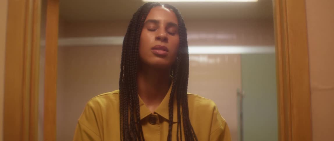 A woman with braids and a yellow shirt stands in a doorway and touches her head while looking at the camera