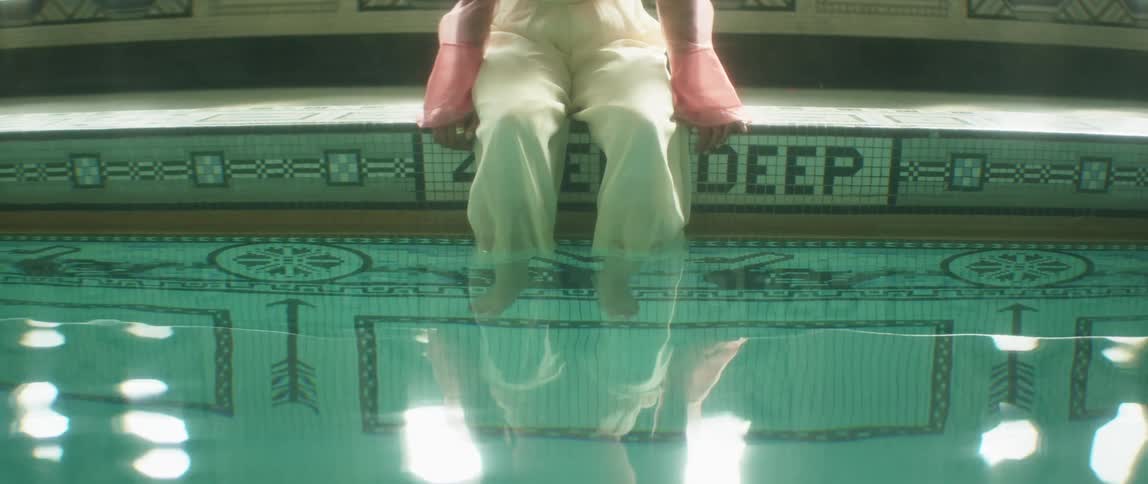 A woman sits on a tiled ledge of a swimming pool with her legs in the water