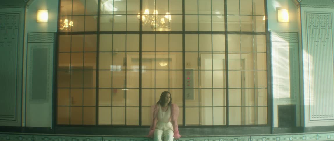 A woman in a pink taffeta blouse sits on a ledge in a building in front of a large window