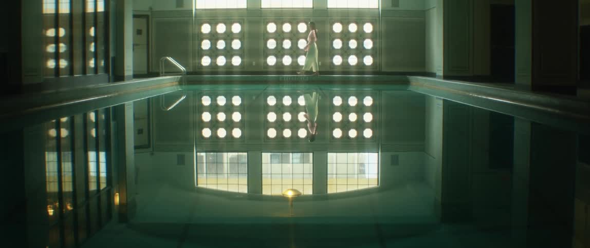 A woman wearing white walks next to an indoor swimming pool with her reflection in the pool