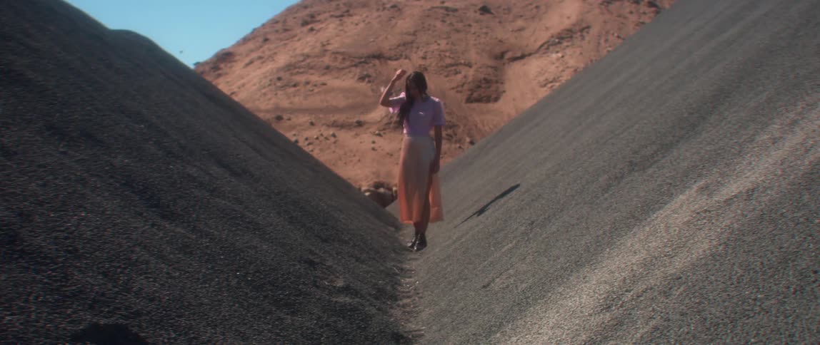 A woman stands outside in the middle of two steep hills with sand dunes behind her as she touches her hair
