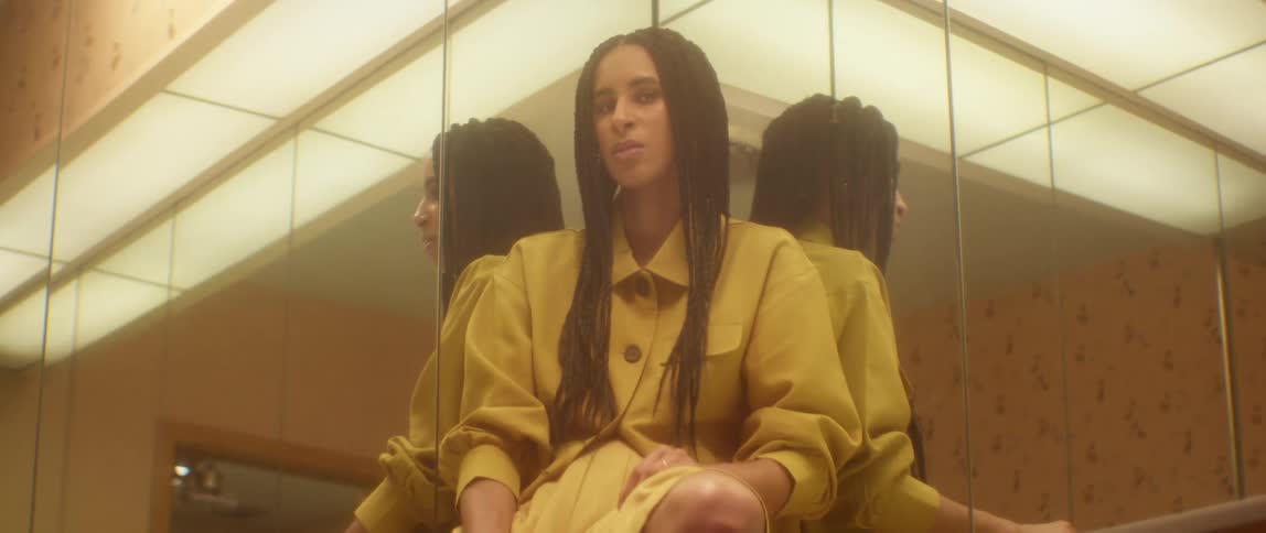 A woman with braided hair sits in a room in a corner with mirrored walls