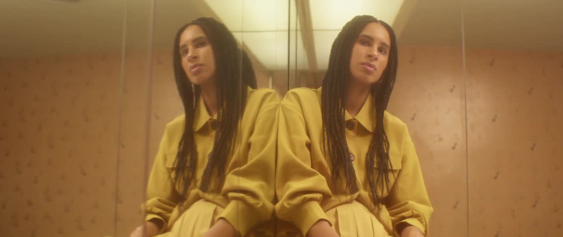 A woman with braided hair and wearing a yellow dress sits in a room indoors with mirrored walls