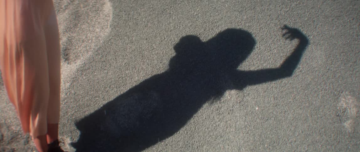 A woman stands on sand moving her arms in dance with her shadow visible on the sand