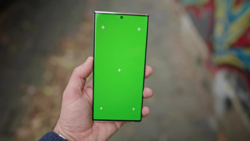 A man is holding a phone with a green screen as he walks in an alley near a building