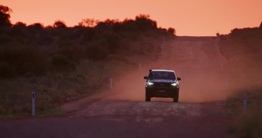 A black truck travels on a dirt road in the rain with an orange sunset sky in the distance