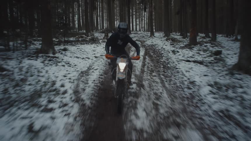 A motorbike speeds through snow covered woods as the rider stands on the pedals traveling on a trail