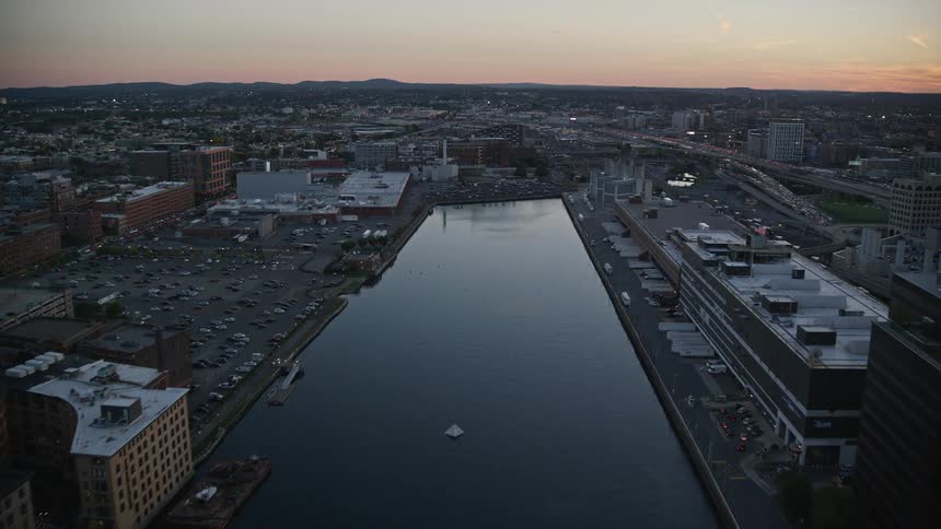 From an aerial perspective the city unfolds with a river at its heart flanked by tall buildings and crossed by a bridge adjacent to the river is a parking lot with cars neatly lined up along the riverbank